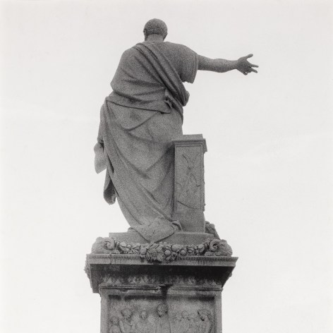 Black and white photo of the back of a stature which depicts a robed man with his arm outstretched. 