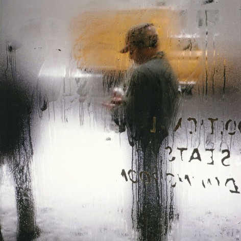 Color photograph of the blurred image of a man on a snowy street, scene through a window dripping with condensation.