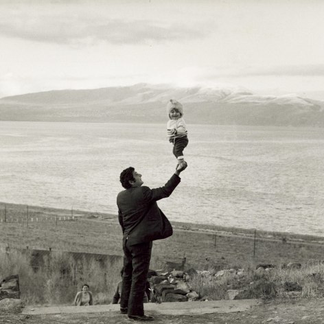 Black and white photo of a man standing in front of a large mountain lake holding a small child aloft with one hand.