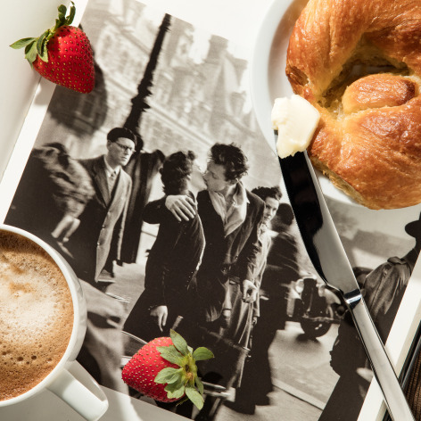 Repriduciton of a Robert Doisneau photograph of a couple kissin with a cup of coffee, a strawberry, and a croissant with butter on top.