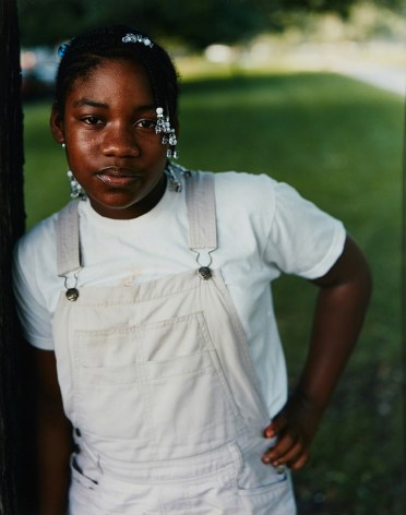 Color photo of a young black girl with beads in her hair, wearing white, leaning against a tree and looking at the camera.