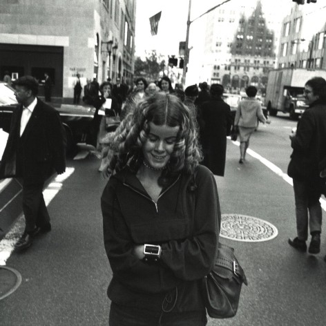 black and white photo shot by Garry Winogrand in 1969 showing a woman smiling and looking down while crossing the street in NYC.
