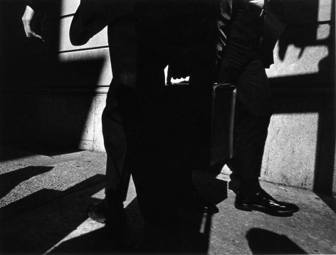 Starkly lit black and white photo showing the leg of pedestrians and a hand holding a briefcase.