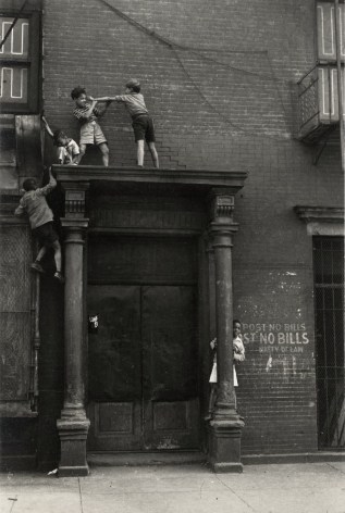 Black and white photo of of young boys who have climbed atop a tall doorway and are now sparring at the top.