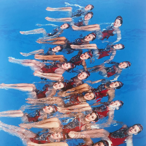 Color photo of a team of synchronized swimmers posing for the camera in a swimming pool.