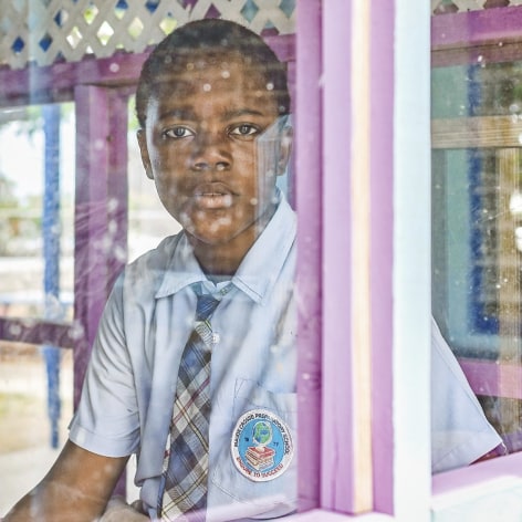 Color photo of a young black boy in a school uniform looking through a window at the camera.