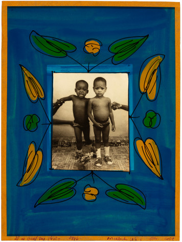 Black and white photo of two very young children, being held up by people outside of the imge, framed in a blue glass frame with hand painted flowers.