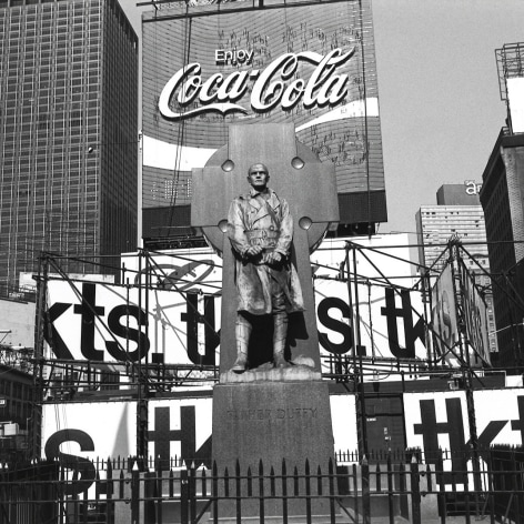 Black and white photo of a bronze statue of military Chaplin Father Duffy, standing in front of a cross, with a hidpodge of urban signing behind him. One prominent sign over his heads reads" " Enjoy Coca-Cola"