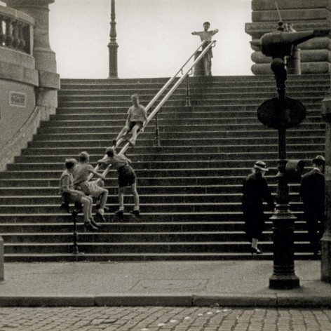 Black and white photo of a group of children watching their friend slide down the handrail on large public stone steps.