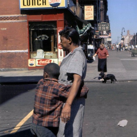 Color photo a 1950s NYC street showing a young boy in. applied shirt embracing a woman who appears to be his mother. A woman walks her dog in the background. 