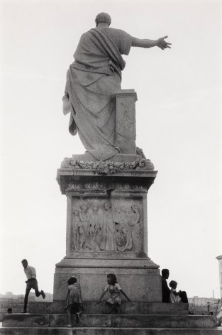 Black and whtie photo of the back of a large statue of a robed man with hand outstretched, with people gathered around the steps at the base.
