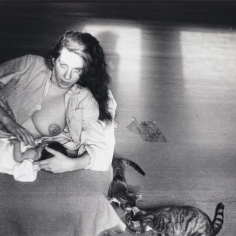 Black and white photo of young mother nursing an infant on the floor in a beam of sunlight next to two young cats eating.