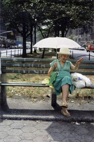 Color photograph of of a smiling woman on a city bench, smiling and waving.