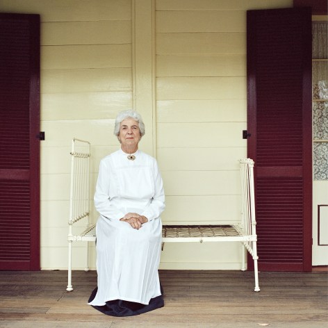 Color photo portrait of a white haired woman in a white dress sitting on an old fashioned bed springs