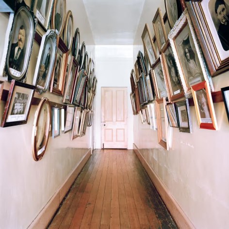 Narrow hallway with wood floors and door at the end, hung with many framed pictures on both sides of the passage.