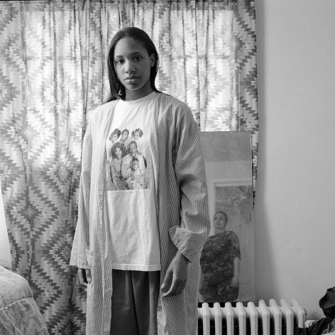 Black and white photo portrait of a young woman looking at the camera wearing an old Cosby Show shirt, in a domestic interior.