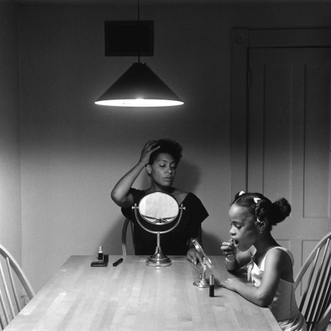 Black and white photo of a mother and daughter sitting at a table, beneath an overhead lamp, putting on makeup while they each look in a mirror.