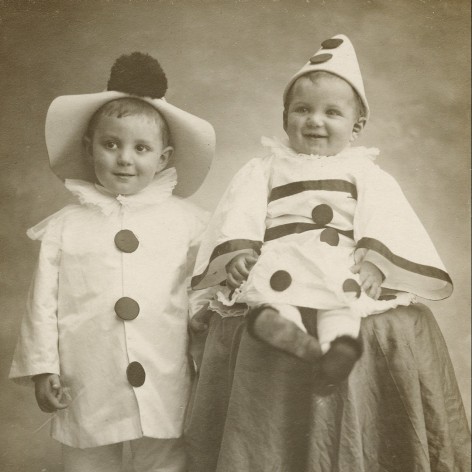 Vintage black and white photo of two young children dressed in clown costumes, posed in a photo studio. 