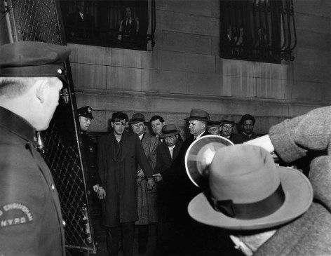 Black and white photo of an arrested man being walked by police in front of news photographer.