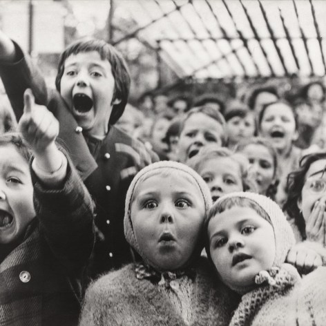 Black and white photograph of children's expressions of surprise and excitement as they watch from the audience of an outside performance. 