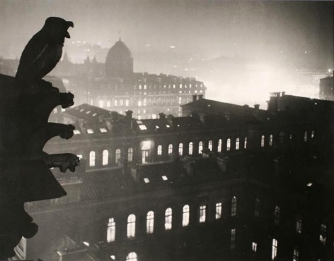 Black and white image. Stone owl and gargoyles atop Notre Dame, silhouetted against Paris at night, shot by Brassaï.