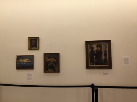 White gallery wall with a triangular cluster of small dark colored paintings on the left and one medium sized dark painting on the right. All of the paintings are in dark brown frames except for the smallest one which is on the top left in a gold frame. From left to right the paintings depict: a female nude figure laying on a white cloth, a female portrait, a man sitting in an armchair, and a young man sculpting.