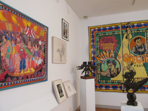 Two white gallery walls intersect to the left of us. On the right wall a colorful tapestry is hanging, and on the left the vibrant composition by Mimi Gross, &quot;Fred Dean's Circus in Maine&quot; is hung up closest to us. Between those two pieces there are four small art works hung in three cream frames and one black frame. A small dark brown sculpture of four female figures, by Chaim Gross, &quot;The Flying Ballerinas&quot; sits in the middle of the two walls and a small, dark brown sculpture of a person standing on a horse is in the bottom right of the frame, in front of the other pieces.
