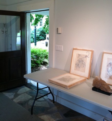 Small, pinkish sculpture and three ink drawings in white and light brown frames in bubble wrap laying on a white fold out table. They are placed in a white room with the door open revealing greenery outside.