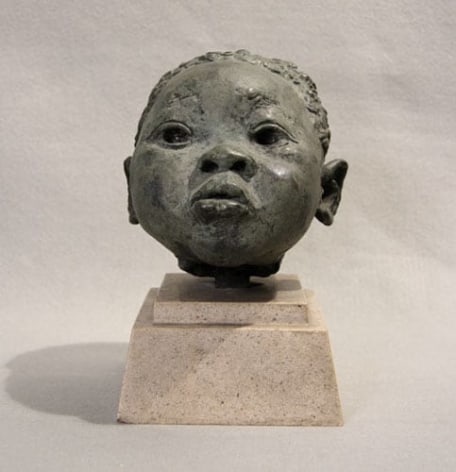 Metal sculpture of the head of a young boy. His head is very round, with large eyes with a wide placement on his face. His noise is barely prominent, with only the circular nostrils and the tip of it standing out. His lips are small and circular and are slightly parted, giving him a confused, concerned, innocent expression.