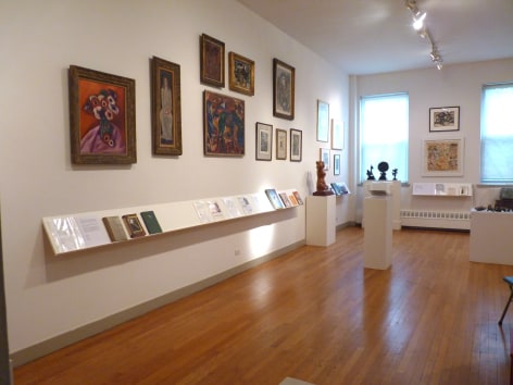 White walled gallery space with light brown wood flooring. Closest to use on the left on a wall going diagonally to the right is the painting by Marsden Hartley, &quot;Anemones&quot;. On the right side of the frame you can see several small sculptures mounted on white rectangular blocks, and beneath the framed art works on the left wall there are pieces of paper and books scattered on a white shelf.