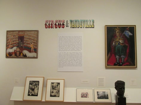 White gallery wall with large printed description beneath a decorative title &quot;Circus &amp; Vaudeville&quot; in colorful, vintage type. On the left of the description is a large painting with performers and an audience in a medium brown frame, and to the right is painting of man wearing a crown and a red cape in a beige frame with a gold outside. On a small shelf beneath the paintings and description there are two smaller artworks in beige frames on the left, and two photos in black frames and a black bust of a man's head on the right.
