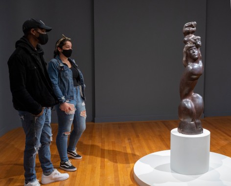 Photo of two individuals (left) viewing I LOVE MY BABY, 1948, which is positioned on a rounded white base. The wooden sculpture depicts a woman with long hair, crouched so that her legs are drawn close to her tall torso. She is supporting the figure of a baby, whose head touches hers but whose body is raised above its head.