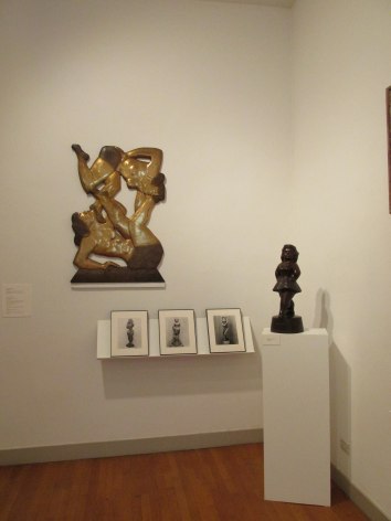Two white gallery walls intersect to the right of us. On the left wall is a yellow-brown sculpture of two female figures balancing on each other hung up with three small black and white photos in black frames on a shelf beneath it. In the corner where the walls intersect is a dark brown, small sculpture of woman stepping forward.