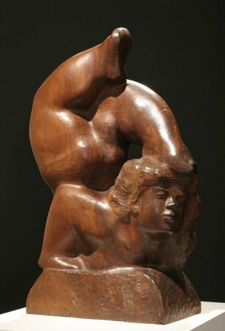 Sculpture of a woman in a deep backbend, legs over her head with her left toe touching the top of her head. The sculpture is carved in a warm, reddish brown. The texture is smooth and shiny except for her hair and the base where she rests her hands. Both areas have a bumpy texture left by the tools of the artist.