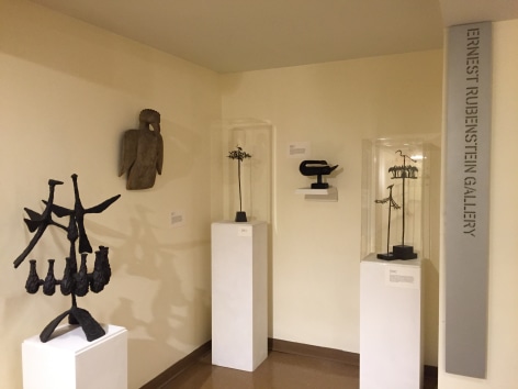 Photo of two cream gallery walls intersecting on the left of us with medium brown flooring beneath them. On the left wall closest to us there is a black sculpture two birds with a series of small vase like forms beneath them mounted on a white rectangular block and a light brown sculpture of a bird hung up. Where the two walls intersect there is a thin, tall sculpture of a tree with sparse leaves and a tiny shape of a bird on the top in a glass box on a rectangular mount. On the right wall there is a black sculpture of an organic, crescent shaped form on a small shelf and two more tree-like sculptures with birds on the top of them in glass boxes with rectangular mounts. On the far right there is a vertical metal sign that says, &quot;Ernest Rubenstein Gallery&quot;.