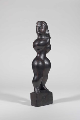 Photo of a sculpture depicting a standing woman in a shiny, dark wood. She is standing on a small pedestal of the same wood and looking over her right shoulder as she clasps her hands together by her left shoulder.
