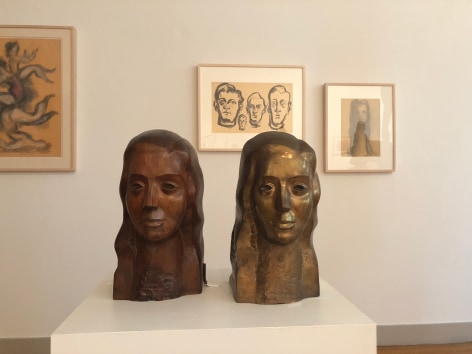 Two identical busts of a woman on a white mount. In the background hung on the wall is a drawing in black ink of of the faces of four men with two other drawings in charcoal, one in the far left corner and the other directly to the right of it.