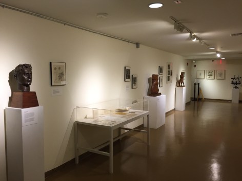 Hallway of a gallery with a long white wall on the left and a small, square back wall visible, medium brown flooring, and a low white ceiling. From left to right along the wall there is a dark brown bust of a man, a glass box with papers and a book inside it on a white table, a medium brown sculpture of a vaguely humanoid form, a medium brown sculpture of unidentifiable, animal like shape, and finally a black sculpture of a tree with birds on it on the back wall. On the walls dotted between these displays are a few pieces of artwork hung up.