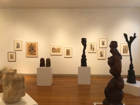 White gallery wall with medium brown wood flooring beneath it. In front of us, there are sculptures from left to right of: a white, short bulbous shape, two female heads, a thin, tall parent bird feeding its children, a tall, large woman carrying her child above her head, and a thin, tree-like, triangular shape. On the wall there are various framed sketches of the sculptures.