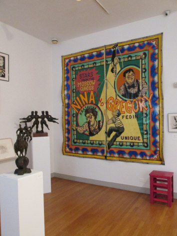 Photo of a white gallery wall with a large, colorful tapestry hung on it. To the left of us there are two small, dark sculptures mounted on rectangular white blocks. On the very bottom right of the frame there is a small red stool beneath the tapestry.