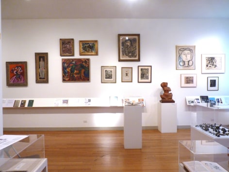 Front view of the wall with framed art works. Closest to us are glass covered box installations, and to the right of us against the wall is small medium brown sculpture of female form seated cross legged.