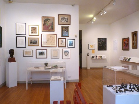 View of the front three walls of the exhibition. The one one the left is the same wall that features the portrait of a young man and is also covered in a collection of framed drawings. In the bottom left corner there is a small sculpture in dark brown of a young man's face. To the right you can see the initial mounted description of the exhibition as well as more artworks on the walls.