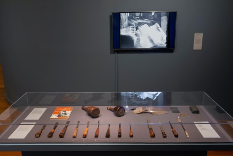 Photo of the encased wooden sculpture tools and equipment featured in the exhibit. On the wall behind the case is a mounted tv playing a video on Chaim Gross' work. On the wall where the mounted tv is, there is a quote from Chaim Gross in white against the faded navy walls.