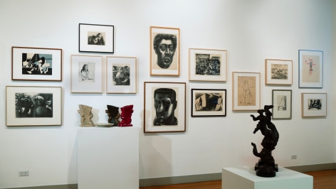 View of an exhibition with many works on the wall in a salon style hang. There are sculptures on pedestals as well.