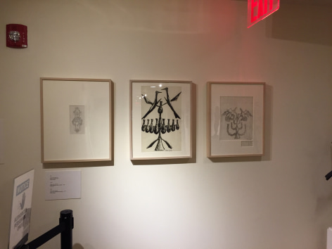 White gallery wall with three similarly sized medium illustrations in beige frames. Each composition depicts drawings of the tree-like stands with birds on the top of them, with the outermost pieces being light sketches and the middle being a larger, black colored composition.
