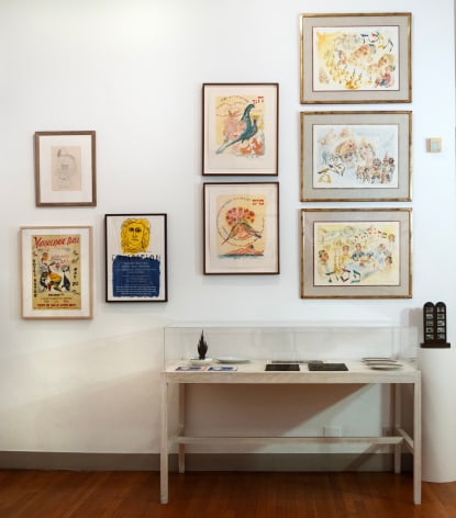 View of a wall in an exhibition with works in a salon style hang. There is a display table in front of the wall with ceramics and two-dimensional works in it as well.