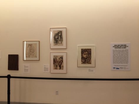 White gallery wall with a black rope fence in front of it. On the right there is a description with the title, &quot;Home Away From Home: Chaim Gross and 70 Years at Educational Alliance&quot;. To the left of the description are three charcoal illustrations of people in light beige and silver frames. In the bottom left there is a black painted canvas or wood piece.