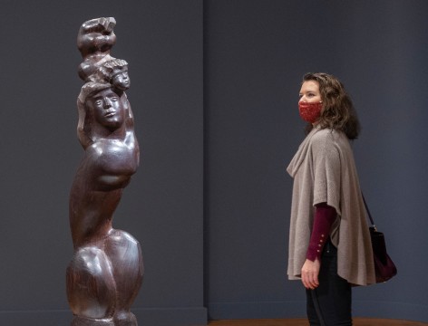 Photo of a masked woman (right) viewing I LOVE MY BABY, 1948, which is positioned on a rounded white base. The wooden sculpture depicts a woman with long hair, crouched so that her legs are drawn close to her tall torso. She is supporting the figure of a baby, whose head touches hers but whose body is raised above its head.