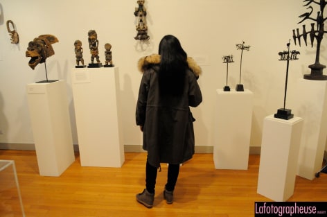 Person with long black hair wearing dark grey winter clothing facing backwards away from us. They are looking at a collection of African Art mounted on white blocks in wood and metal. There are also two other small pieces of wood art mounted on the wall.