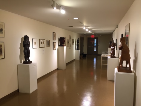 Hallway of a gallery with white walls and medium brown floors. The closest piece of artwork to us on the left wall is a medium black sculpture of a humanoid figure, in the middle is a medium brown bust of a man, and furthest away is a medium black sculpture of two thin, organic shapes stacked vertically on top of eachother. On the right wall closest to us is a mid-sized medium brown sculpture of a person sitting on an unidentifiable organic shape, in the middle is a darker brown, smaller sculpture of a female form, and furthest away is a dark brown bust of a man. Between the sculptures there are various framed artworks hung up on the walls.